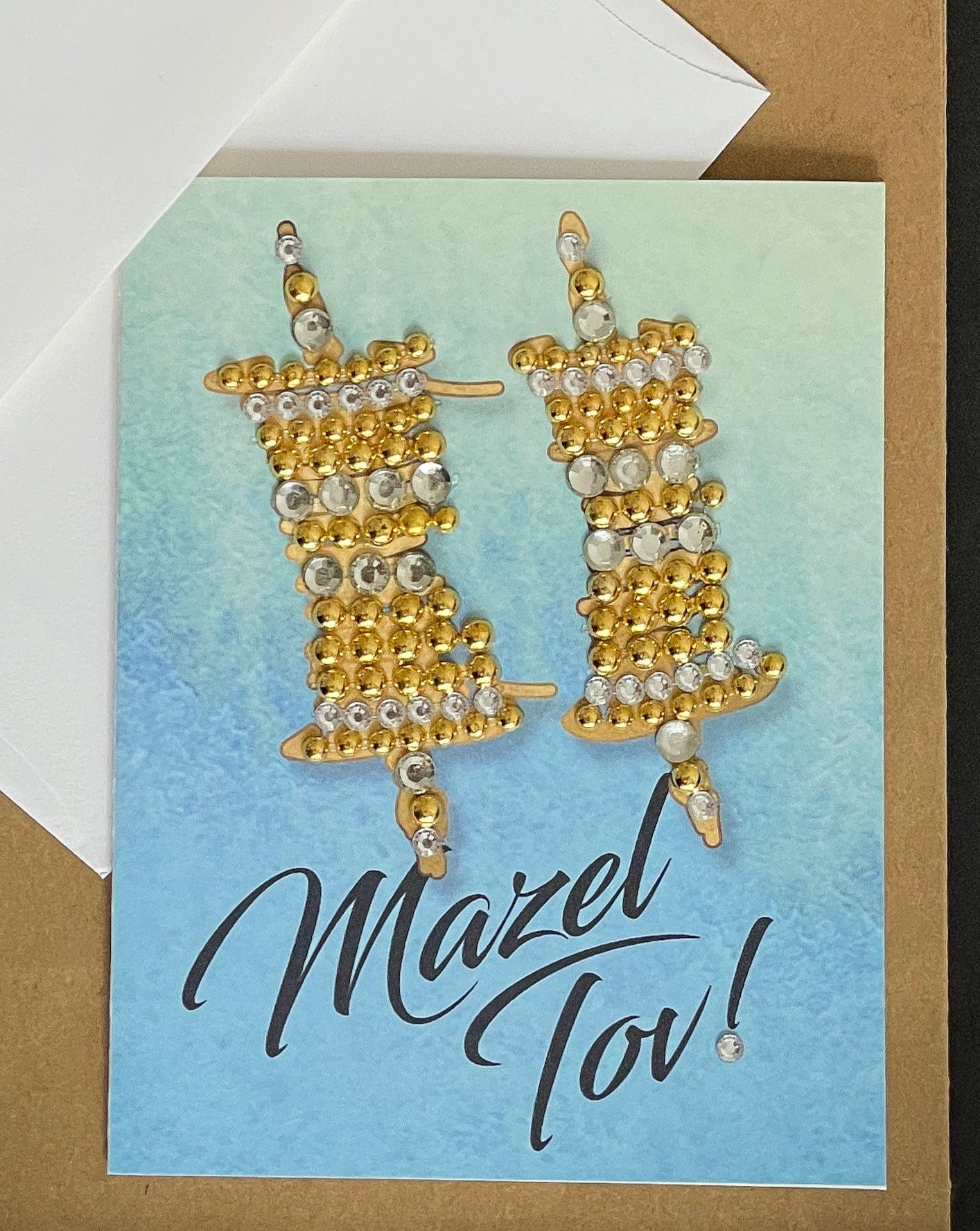 this is a note card on a craft paper gift bag with a white envelope and a black marker , the card says Mazel Tov with a Gold Torah Scroll on Blue Watercolour Background