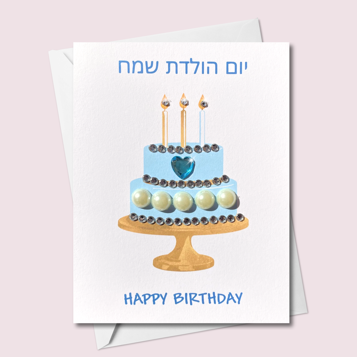 Hebrew and English happy birthday card featuring a blue two-tier cake with sparkling jewels, pearl-style accents, a blue heart and three candles on a gold cake stand.