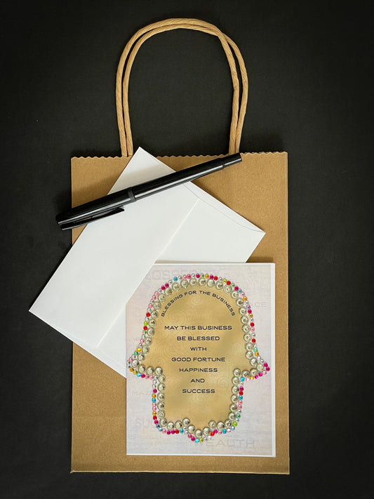 this is a note card on a craft paper gift bag with a white envelope and a black marker gold hamsa facing down with the business blessing written in english outlined in multicolour jewels