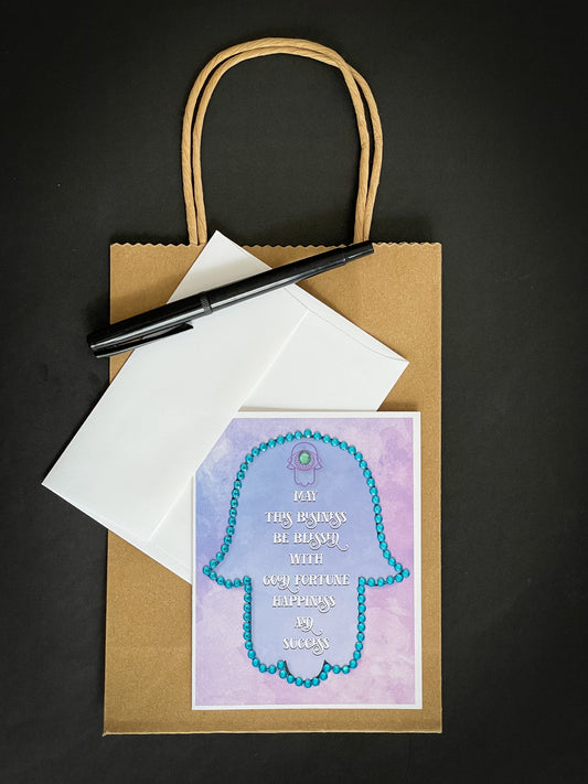 this is a note card on a craft paper gift bag with a white envelope and a black marker purple hamsa facing down with the business blessing written in english outlined in blue stones