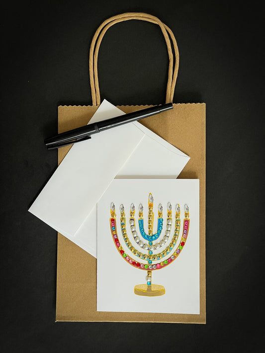 this is a note card on a craft paper gift bag with a white envelope and a black marker , the card has a jewelled menorah or Chanukiah 