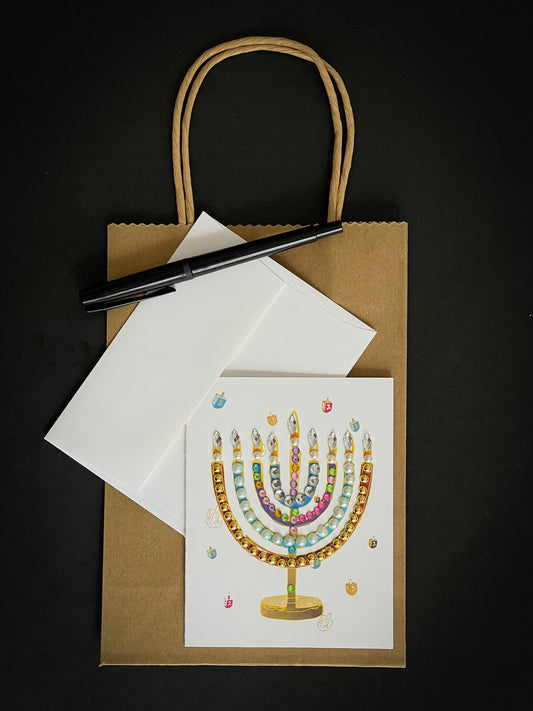 this is a note card on a craft paper gift bag with a white envelope and a black marker , the card has a jewelled menorah or Chanukiah  with mini driedels in the background
