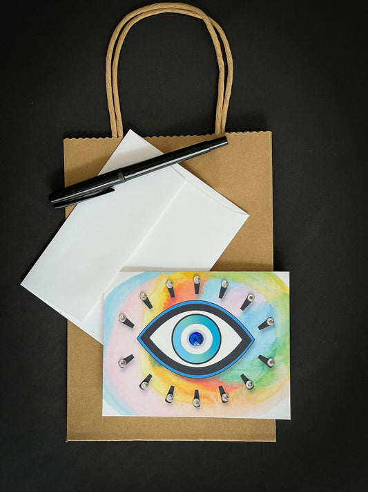 this is a note card on a craft paper gift bag with a white envelope and a black marker , the card has an evil eye symbol on a colourful background