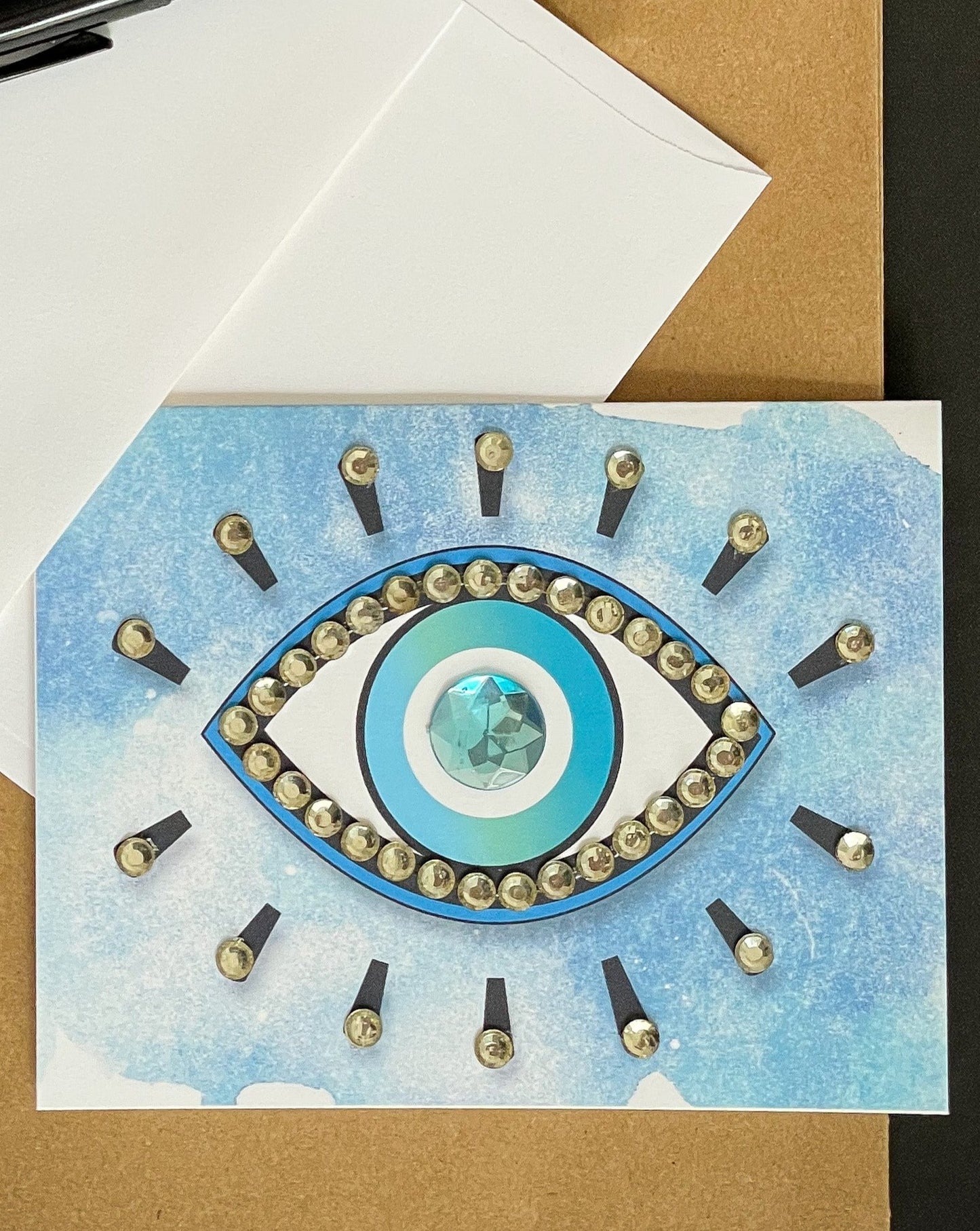 this is a note card on a craft paper gift bag with a white envelope and a black marker , the card has an evil eye symbol on a blue background