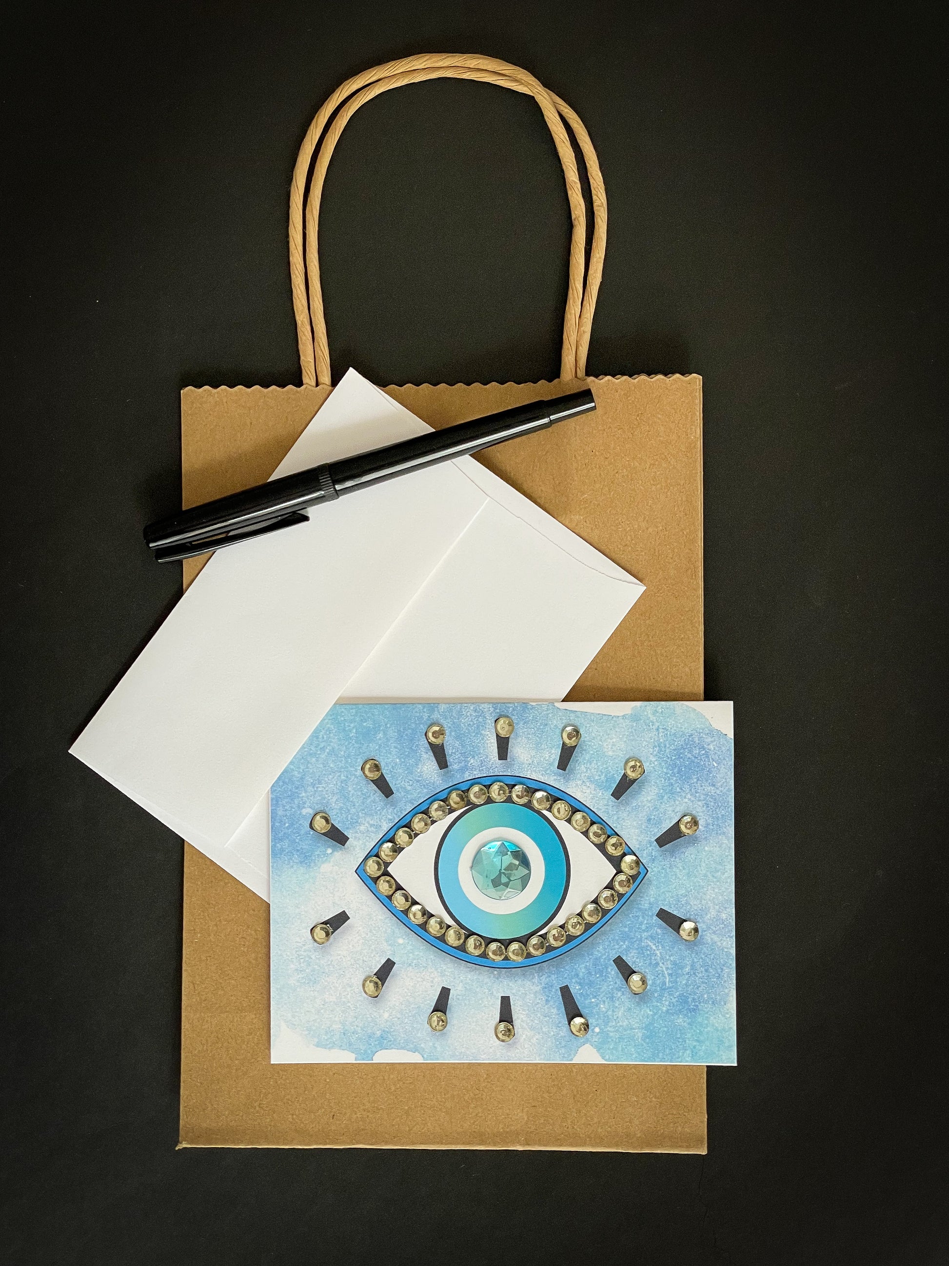 this is a note card on a craft paper gift bag with a white envelope and a black marker , the card has an evil eye symbol on a blue background