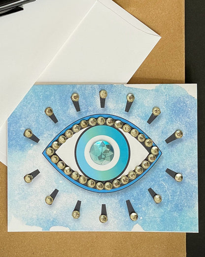 this is a note card on a craft paper gift bag with a white envelope and a black marker , the card has an evil eye symbol on a blue background