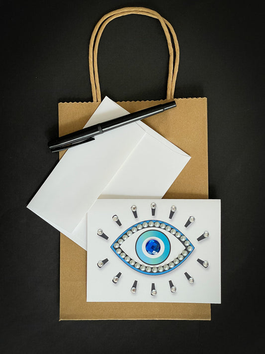 this is a note card on a craft paper gift bag with a white envelope and a black marker , the card has an evil eye symbol decorated with pearls and clear stones