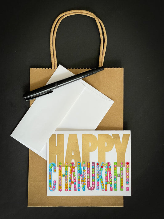 this is a note card on a craft paper gift bag with a white envelope and a black marker , the card has Happy Chanukah written on it 