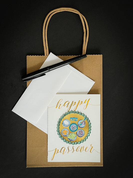 this is a note card on a craft paper gift bag with a white envelope and a black marker , the card has Happy Passover with a decorated gold Sedar plate