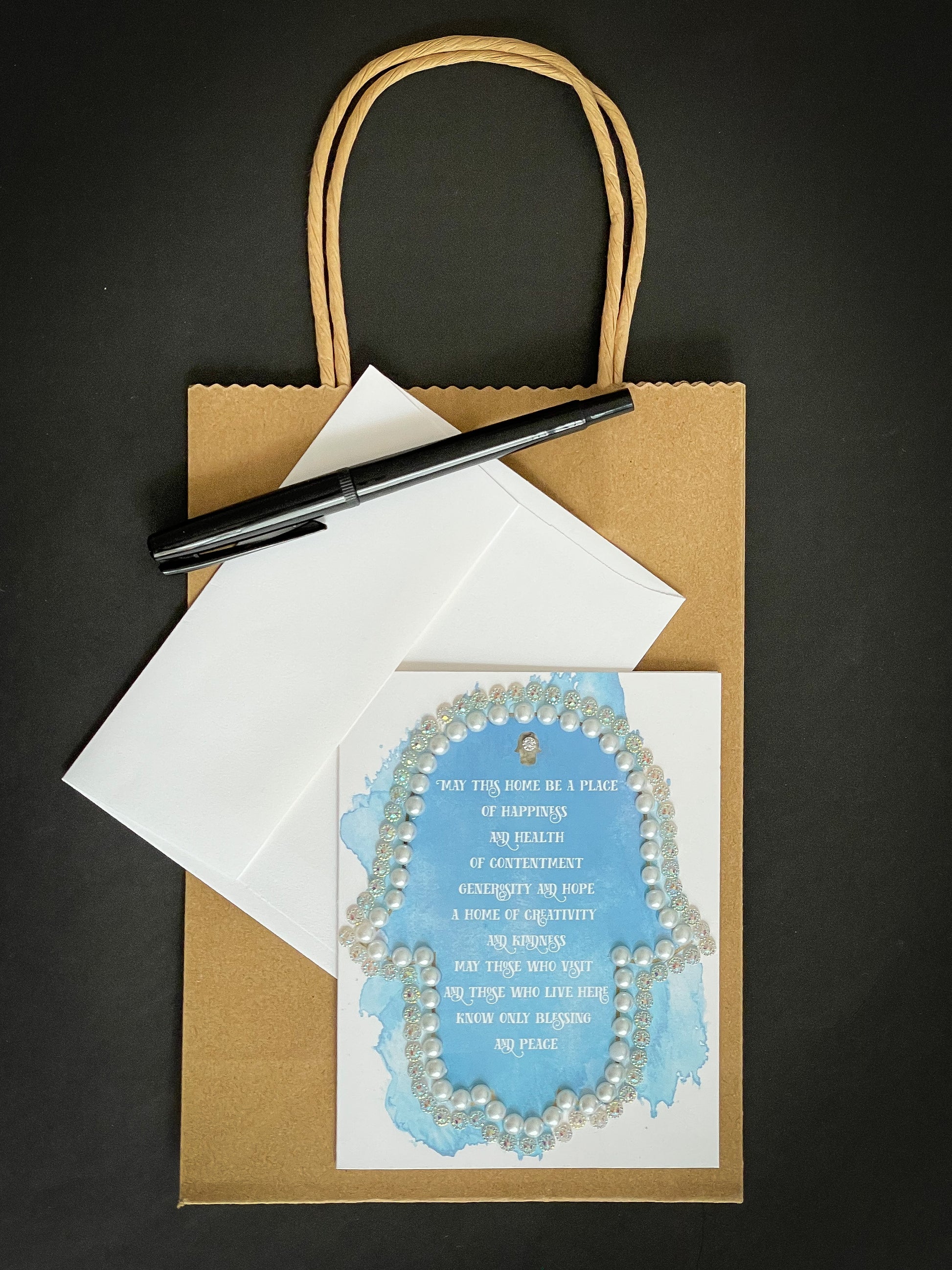 this is a note card on a craft paper gift bag with a white envelope and a black marker , the card has a hamsa with the traditional Jewish home blessing on a blue watercolour background decorated by pearls and jewels 