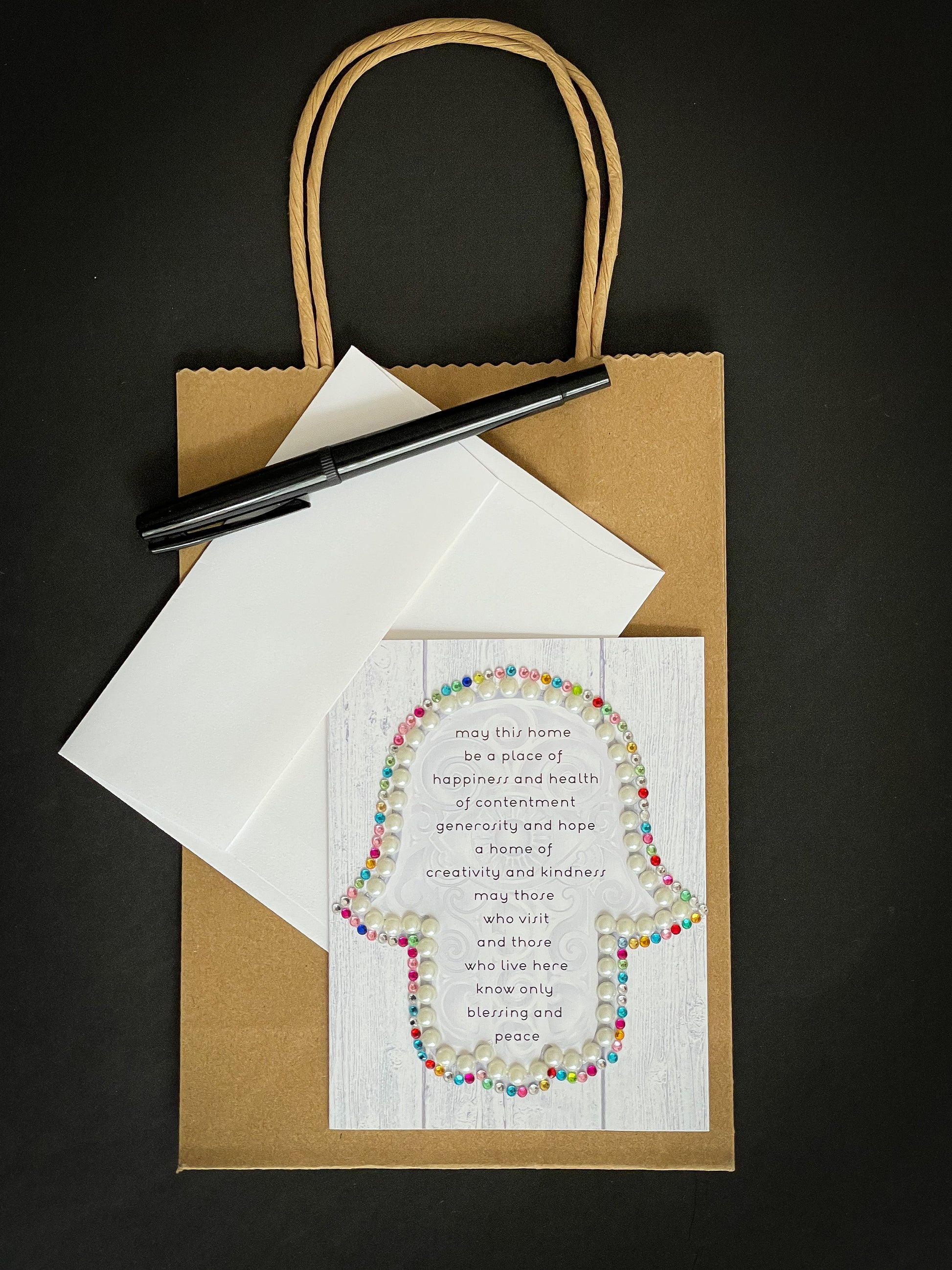 this is a note card on a craft paper gift bag with a white envelope and a black marker , the card has a hamsa with the traditional Jewish home blessing on a light wood background decorated by pearls and jewels 