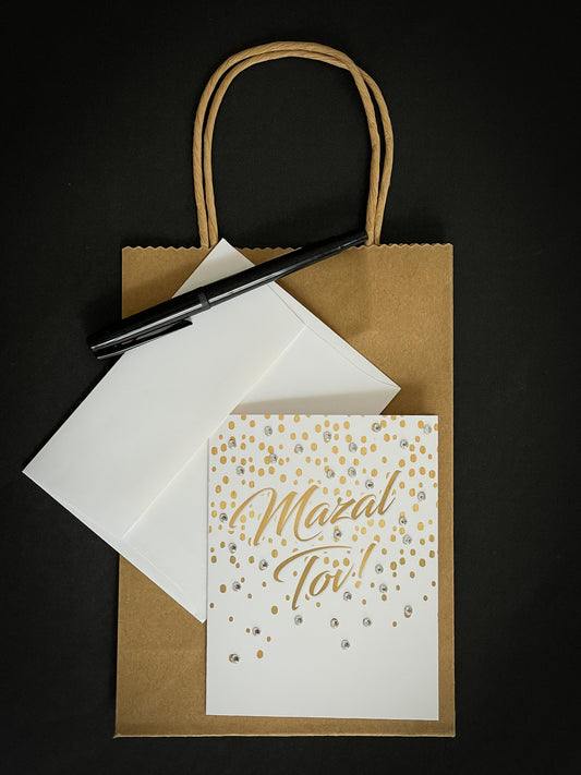 this is a note card on a craft paper gift bag with a white envelope and a black marker , the card has mazal tov written on it with gold confetti 
