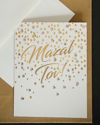 this is a note card on a craft paper gift bag with a white envelope and a black marker , the card has mazal tov written on it with gold confetti 
