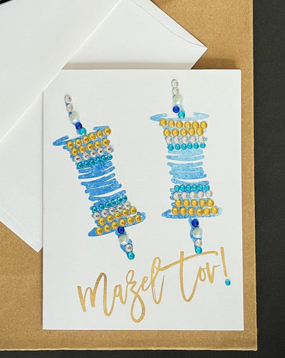 this is a note card on a craft paper gift bag with a white envelope and a black marker , the card says Mazel Tov and has a blue Torah scroll