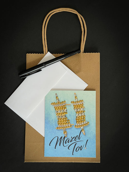 this is a note card on a craft paper gift bag with a white envelope and a black marker , the card says Mazel Tov with a Gold Torah Scroll on Blue Watercolour Background