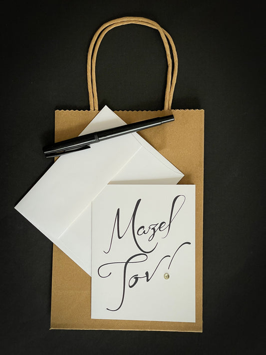 this is a note card on a craft paper gift bag with a white envelope and a black marker , the card says Mazel Tov in black 