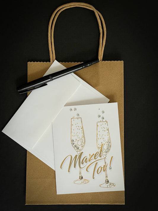 this is a note card on a craft paper gift bag with a white envelope and a black marker , the card says Mazel Tov in gold with champagne flutes 
