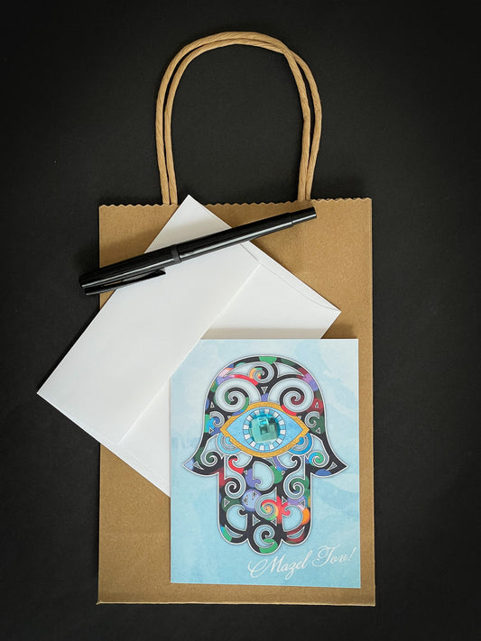 this is a note card on a craft paper gift bag with a white envelope and a black marker , the card has a hamsa on it that looks like stained glass with Mazel Tov written in the bottom right corner