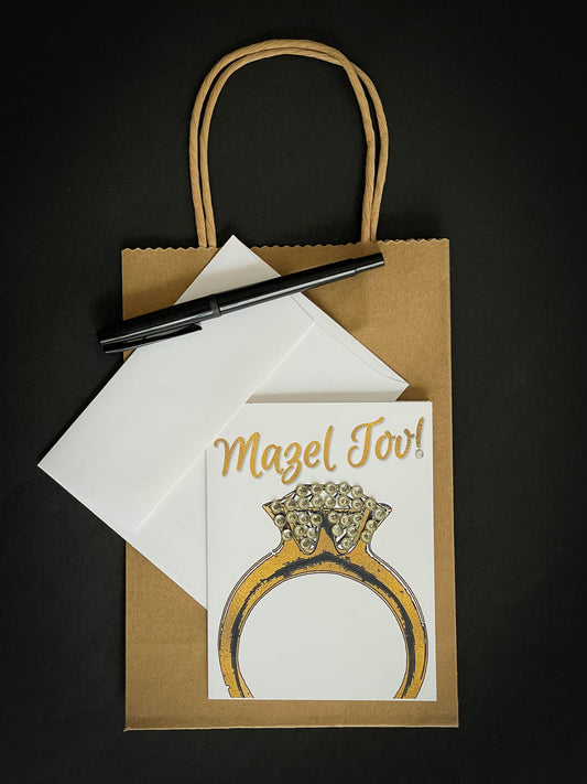 this is a note card on a craft paper gift bag with a white envelope and a black marker , the card says Mazel Tov and has a beautiful engagement ring in gold with jewelled diamond area