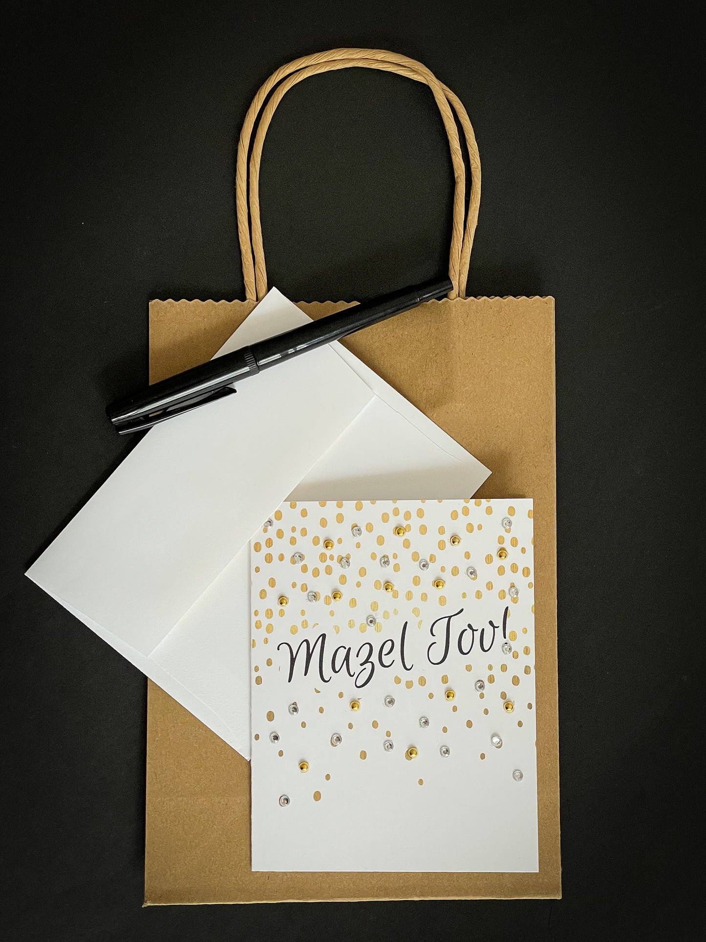 this is a note card on a craft paper gift bag with a white envelope and a black marker , the card says Mazel Tov in black and surrounded by gold confetti design