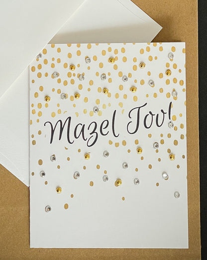 this is a note card on a craft paper gift bag with a white envelope and a black marker , the card says Mazel Tov in black and surrounded by gold confetti design