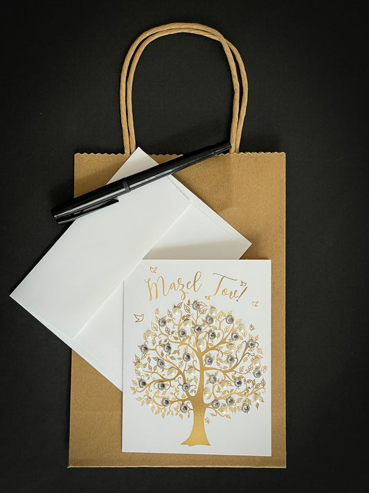 this is a note card on a craft paper gift bag with a white envelope and a black marker , the card says Mazel Tov and there is a gold tree of life