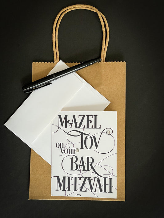 this is a note card on a craft paper gift bag with a white envelope and a black marker , the card says Mazel Tov on your bar mitzvah in a black script font