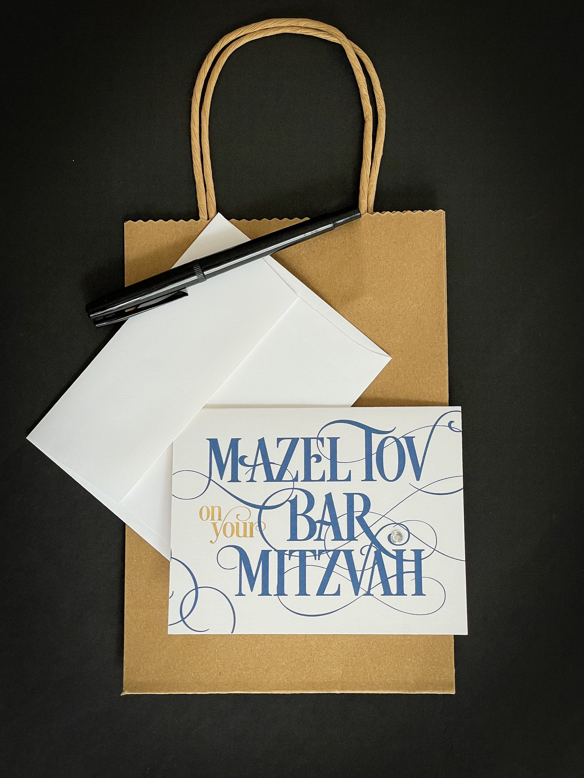 this is a note card on a craft paper gift bag with a white envelope and a black marker , the card says Mazel Tov on your bar mitzvah in a blue script font