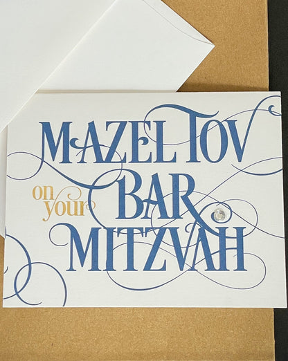 this is a note card on a craft paper gift bag with a white envelope and a black marker , the card says Mazel Tov on your bar mitzvah in a blue script font