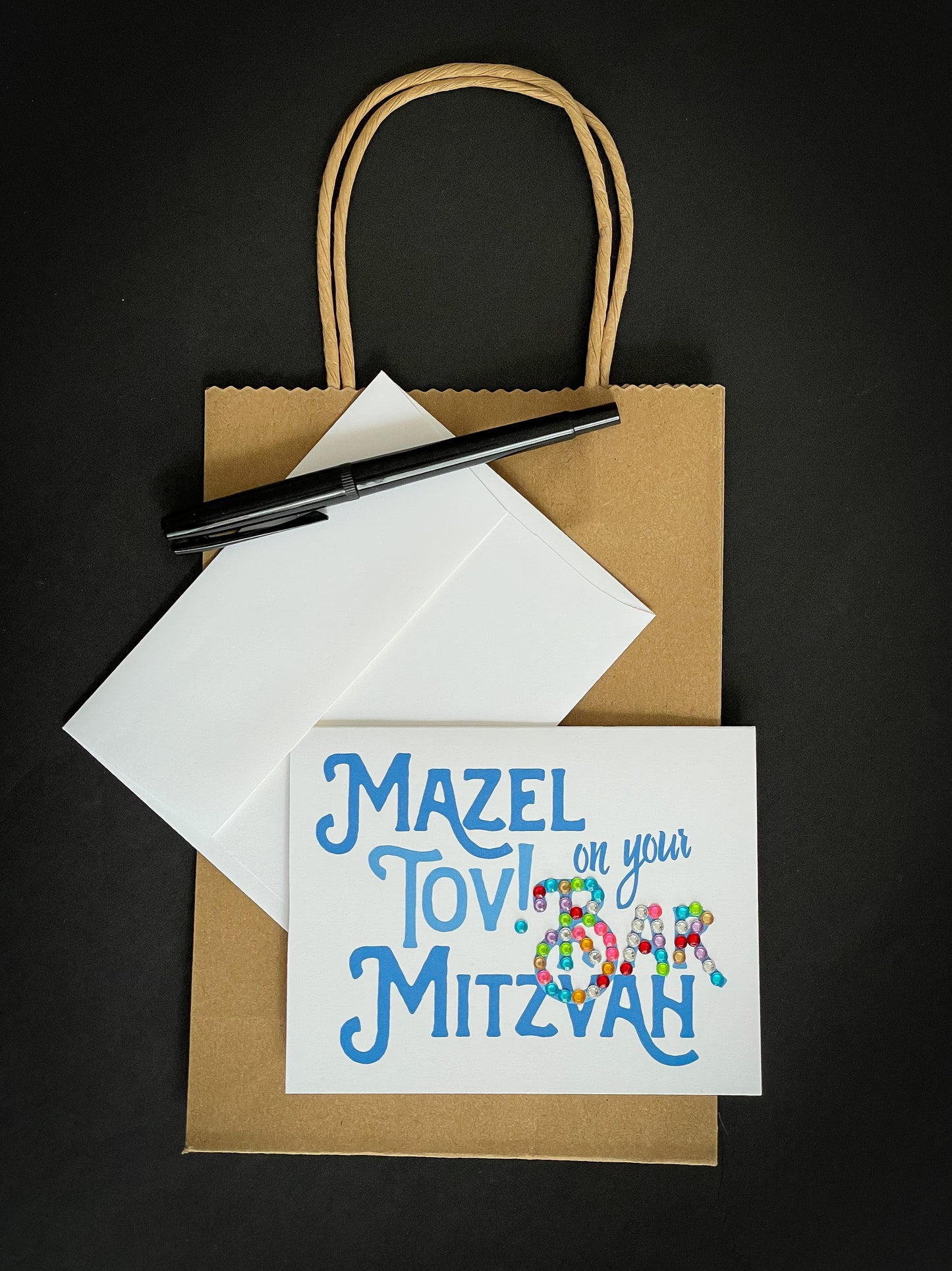 this is a note card on a craft paper gift bag with a white envelope and a black marker , the card says Mazel Tov on your bar mitzvah in blue