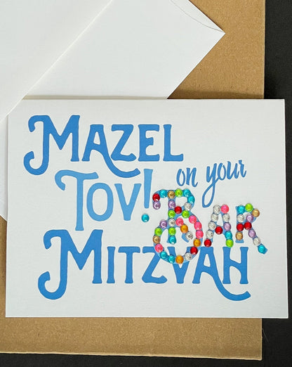 this is a note card on a craft paper gift bag with a white envelope and a black marker , the card says Mazel Tov on your bar mitzvah in blue