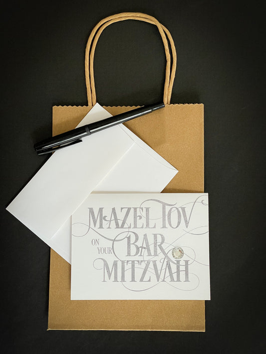 this is a note card on a craft paper gift bag with a white envelope and a black marker , the card says Mazel Tov on your bar mitzvah in a silver script font