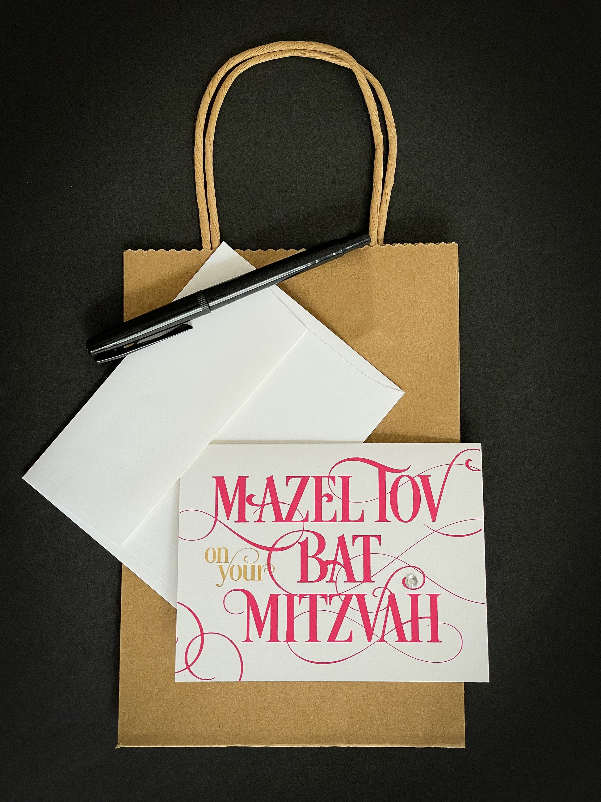 this is a note card on a craft paper gift bag with a white envelope and a black marker , the card says Mazel Tov on your bat mitzvah in a hot pink script font
