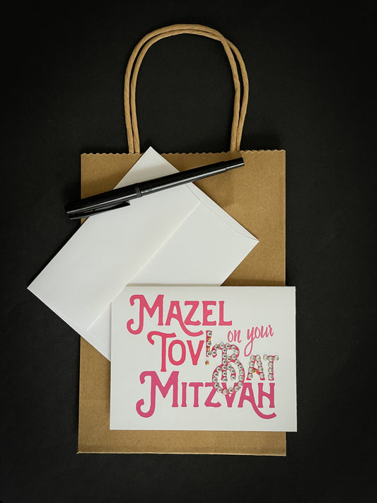 this is a note card on a craft paper gift bag with a white envelope and a black marker , the card says Mazel Tov on your bat mitzvah in pink