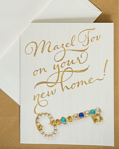 this is a note card on a craft paper gift bag with a white envelope and a black marker , the card says Mazel Tov on your new home