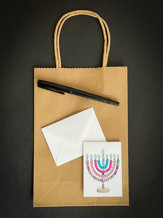 this is a mini note card on a craft paper gift bag with a white envelope and a black marker , the card has a decorated menorah