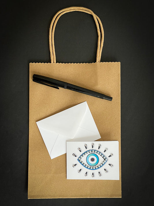 this is a mini note card on a craft paper gift bag with a white envelope and a black marker , the card has an evil eye symbol decorated with pearls and clear jewels