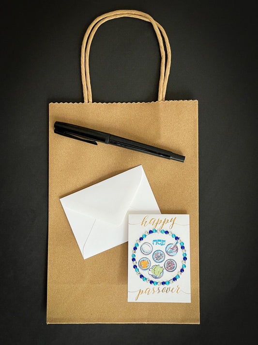 this is a mini note card on a craft paper gift bag with a white envelope and a black marker , the card has Happy Passover with a decorated gold Sedar plate