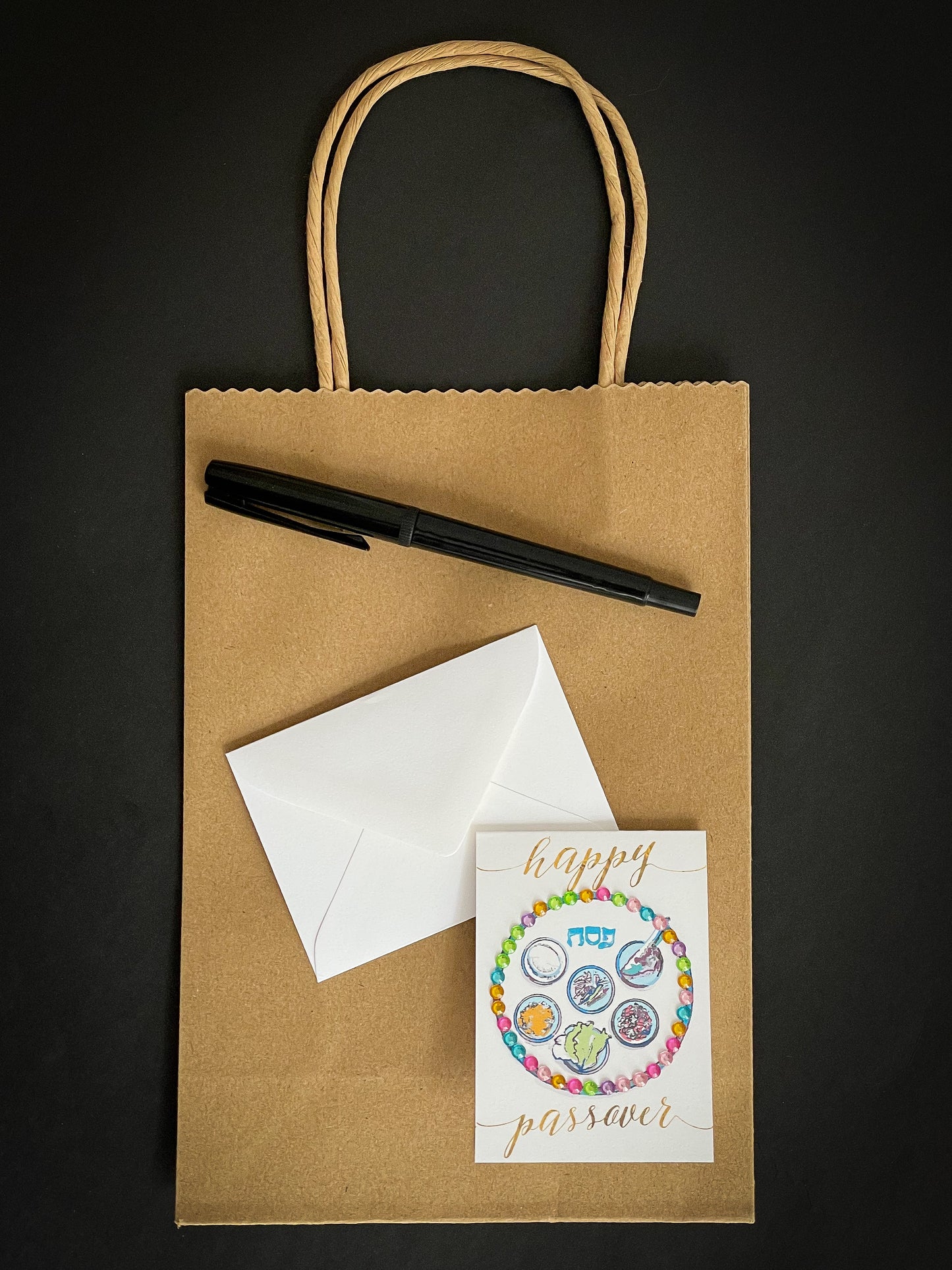 this is a mini note card on a craft paper gift bag with a white envelope and a black marker , the card has Happy Passover with a decorated gold Sedar plate