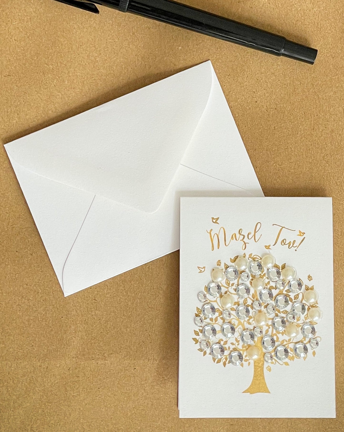 this is a mini note card on a craft paper gift bag with a white envelope and a black marker , the card says Mazel Tov with a decorated tree of life in gold