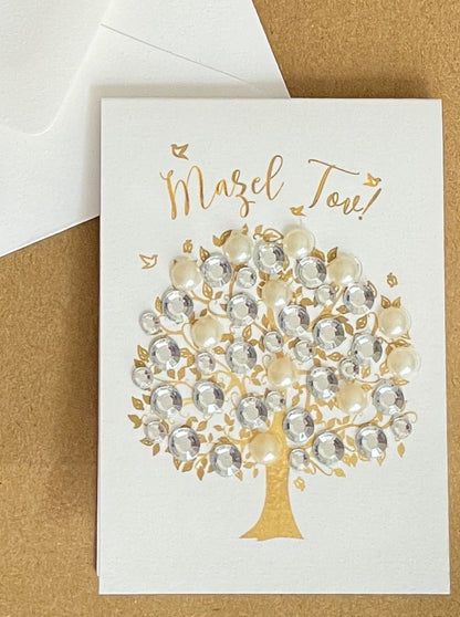 this is a mini note card on a craft paper gift bag with a white envelope and a black marker , the card says Mazel Tov with a decorated tree of life in gold