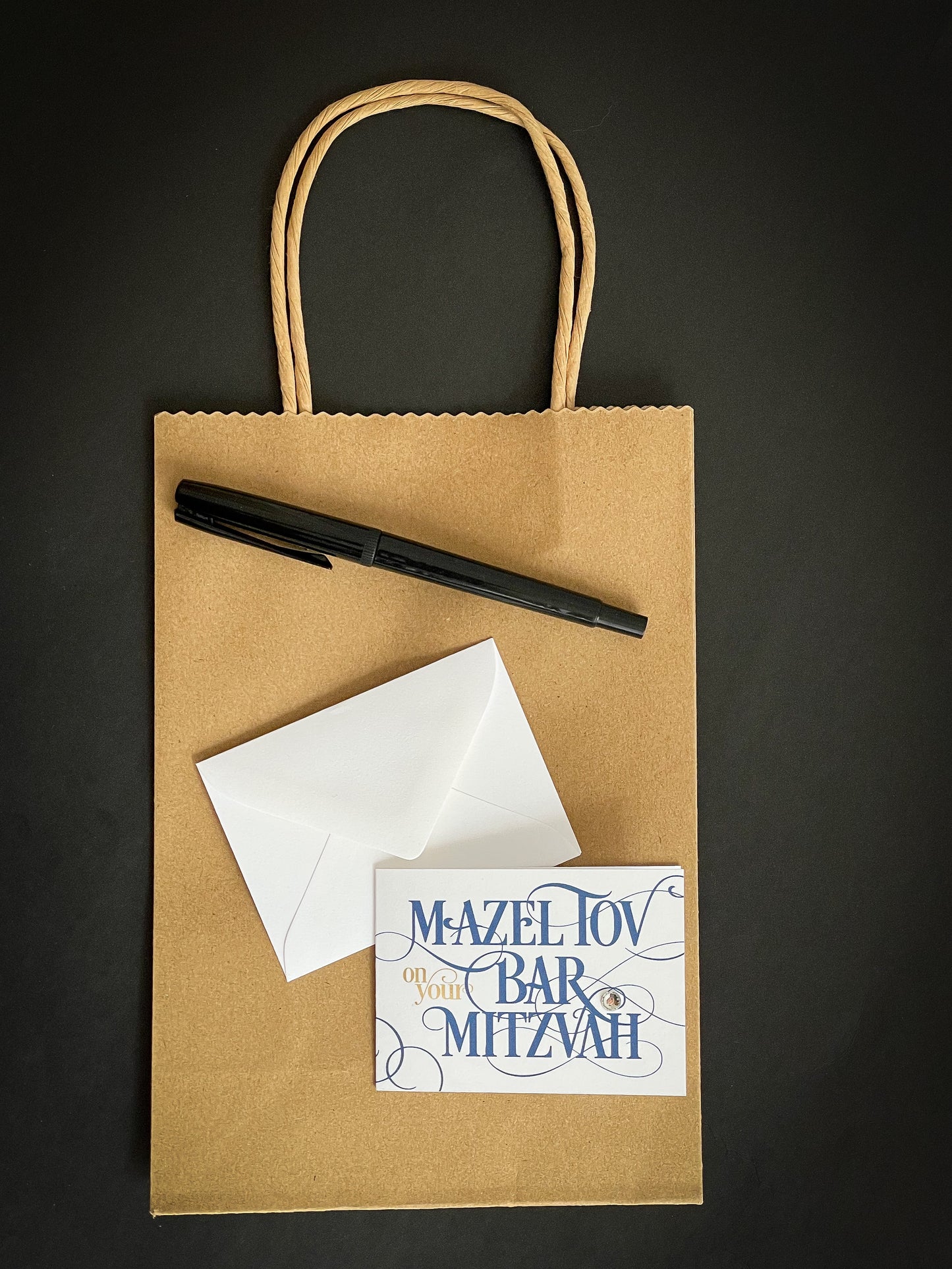 this is a mini note card on a craft paper gift bag with a white envelope and a black marker , the card says Mazel Tov on your bar mitzvah in blue