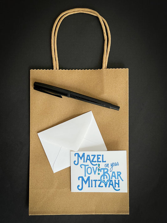 this is a mini note card on a craft paper gift bag with a white envelope and a black marker , the card says Mazel Tov on your bar mitzvah in blue