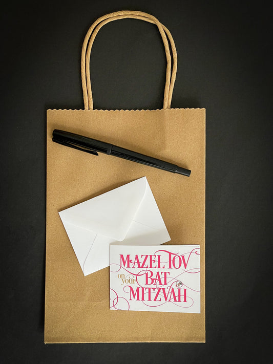 this is a mini note card on a craft paper gift bag with a white envelope and a black marker , the card says Mazel Tov on your bat mitzvah in hot pink