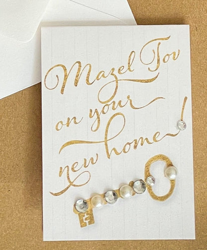 this is a mini note card on a craft paper gift bag with a white envelope and a black marker , the card says Mazel Tov on your new home with a decorated key in gold