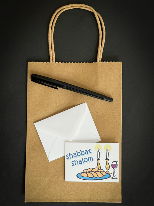 this is a mini note card on a craft paper gift bag with a white envelope and a black marker , the card says Shabbat Shalom with a traditional shabbat set up of challah, wine and candles