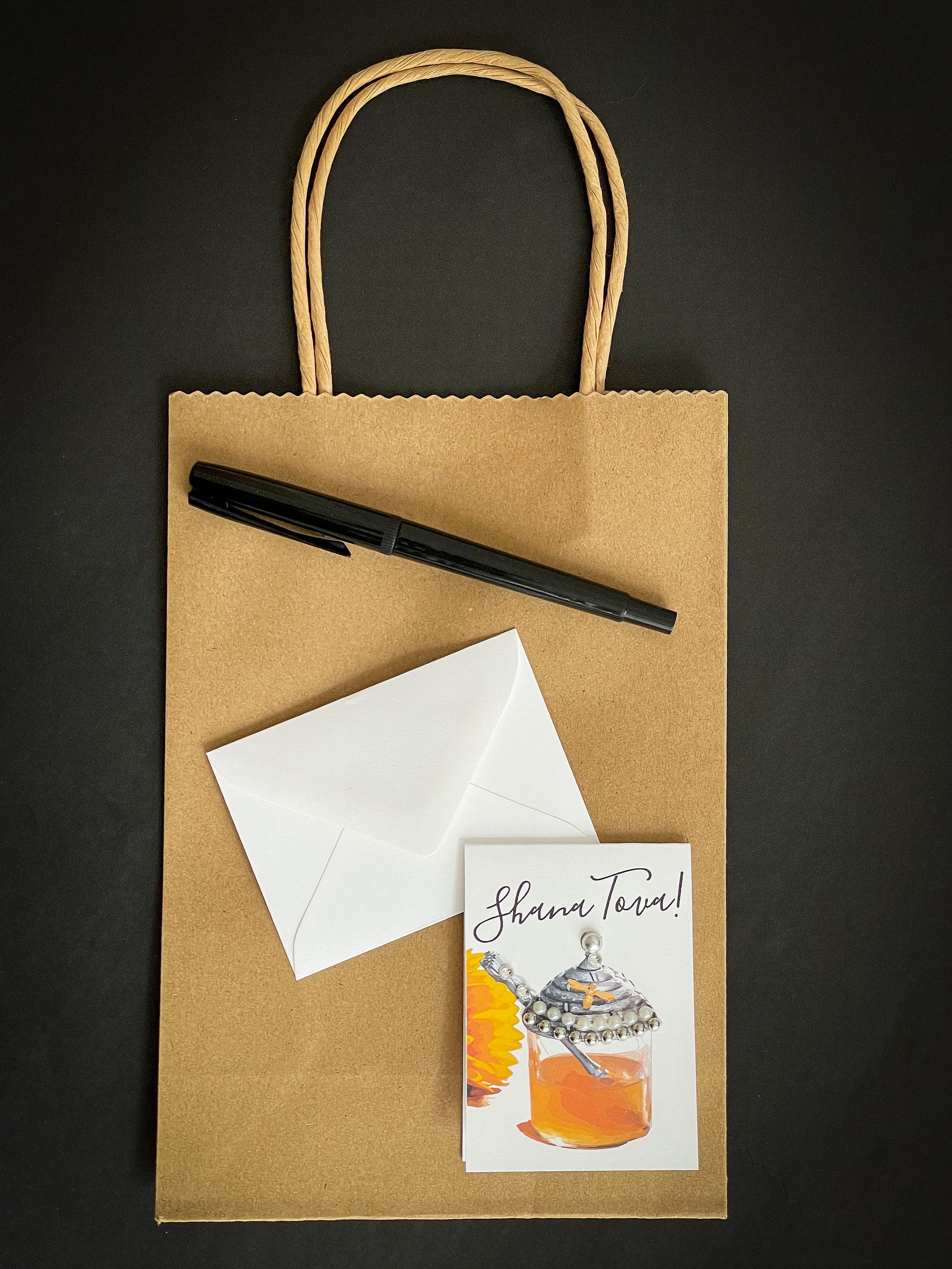 this is a mini note card on a craft paper gift bag with a white envelope and a black marker , the card says Shana Tova in English with a glass jar of honey 