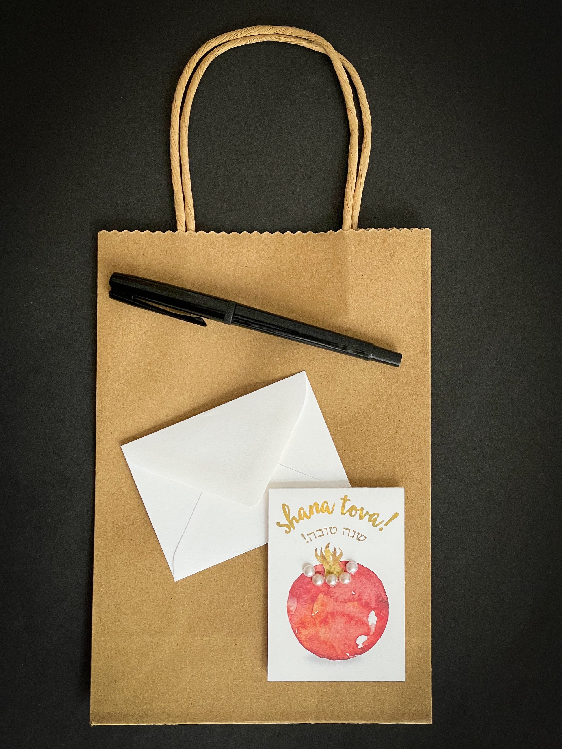 this is a mini note card on a craft paper gift bag with a white envelope and a black marker , the card says Shana Tova in English with a pomegranate