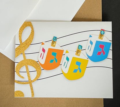 this is a note card on a craft paper gift bag with a white envelope and a black marker , the card has three dreidels and a music note