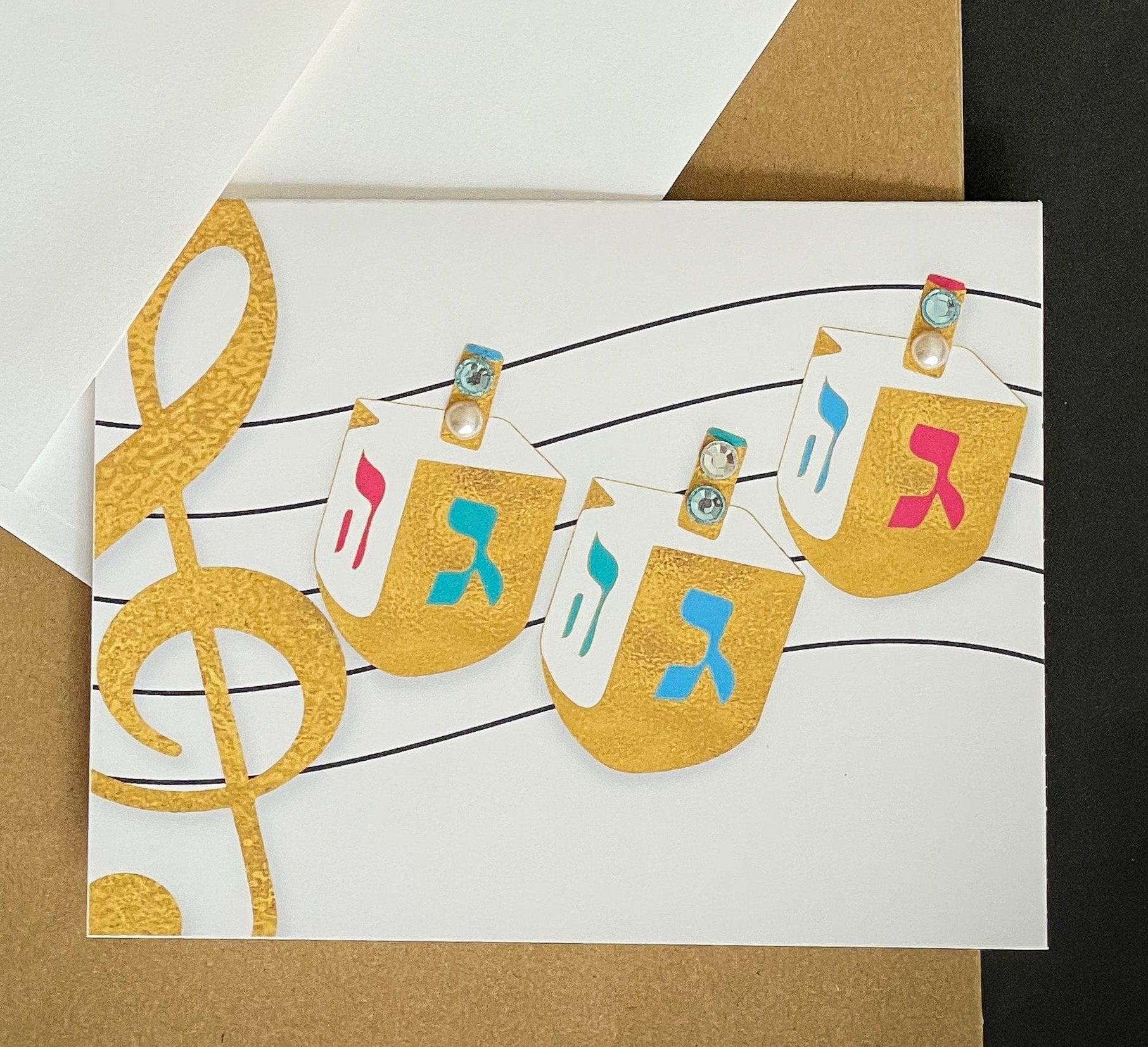 this is a note card on a craft paper gift bag with a white envelope and a black marker , the card has three dreidels and a music note